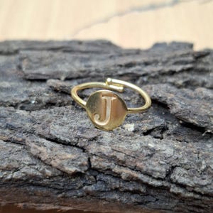 May include: A gold-coloured adjustable ring with the letter "J" on a circular face. The ring sits on a textured, dark brown surface. The ring's band is thin and delicate.