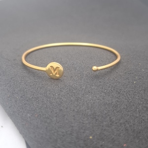 May include: A gold-colored open cuff bracelet. The bracelet features a circular charm with the letter "M" in the center. The bracelet is made of a thin, smooth metal and has a simple, elegant design. The bracelet is shown on a dark gray surface.