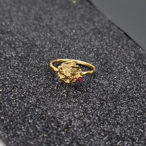 May include: A gold-colored ring with a textured, organic design. The ring features a central, textured element and a small, red gemstone. The ring is set against a dark, glittery surface.