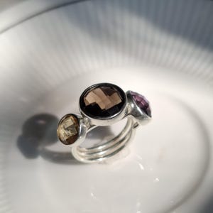 Op de afbeelding: Een zilveren ring met drie ronde, gefacetteerde edelstenen op een rij. De middelste steen is een grote, donkerbruine rookkwarts, geflankeerd door een kleinere goudgele citrien aan de ene kant en een kleinere paarse amethist aan de andere kant.