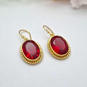 May include: A pair of gold-tone drop earrings featuring oval-shaped, faceted red stones. Each stone is set within a twisted rope-style metal border and hangs from a simple curved ear wire hook, creating a classic and symmetrical jewelry design.