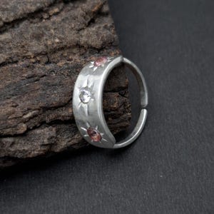 May include: A silver-toned ring with a hammered texture featuring three circular stones set in star-shaped engravings. The center stone is clear, while the two flanking stones are pink. The band has an open design at the back for adjustment.