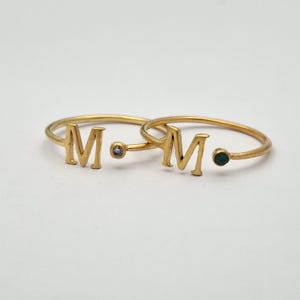 May include: Two gold-colored rings, each featuring the letter "M". One ring has a small, clear gemstone, and the other has a small, green gemstone. The rings are set against a white background.