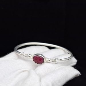 May include: A silver bangle bracelet featuring a central oval-shaped, faceted deep red gemstone. The bracelet has a smooth, polished silver band with a hook-and-eye style clasp mechanism on either side of the gemstone setting.