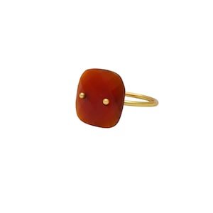 Può includere: Un anello d'oro con una grande gemma rosso-arancio sfaccettata. Due piccoli accenti dorati sono incastonati nella gemma. L'anello ha una semplice e sottile fascia d'oro.