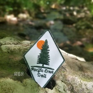 May include: A white diamond-shaped patch with a green pine tree and an orange sun. The text "Pacific Crest Trail" is printed below the tree.