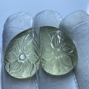 Puede incluir: Dos gemas en forma de pera, de color verde pálido, con intrincados grabados florales. Las gemas se muestran sobre un fondo de tela blanca, mostrando la detallada artesanía y la calidad translúcida de las piedras. Los grabados presentan un diseño floral central rodeado de hojas detalladas.