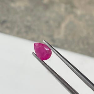 Könnte beinhalten: Ein birnenförmiger, leuchtend rosa Rubin-Edelstein wird von einer silbernen Pinzette gehalten. Der Edelstein hat eine glatte, polierte Oberfläche und eine leichte Transluzenz. Der Hintergrund ist ein weiches, neutrales Grau und Weiß.