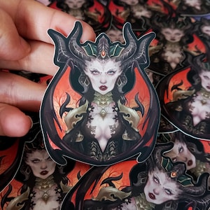May include: A sticker depicting a detailed illustration of a female figure with dark horns, pale skin, and a stern expression. The artwork has a gothic fantasy theme, with a dark colour palette and intricate details. The sticker is held by a hand.