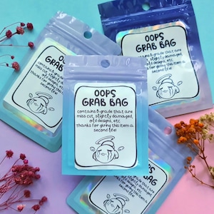 May include: Three clear plastic bags with iridescent holographic designs. Each bag contains a white label with black text that reads "Oops Grab Bag" and a cartoon illustration of an angel with a sad expression. The text on the label explains that the bag contains B-grade items that are slightly damaged, miss cut, or have old designs.