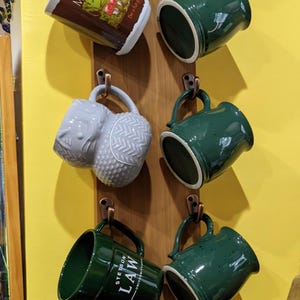 May include: A wooden mug rack displaying various ceramic mugs. The mugs are in shades of green, white, and brown, with unique designs and text. Some mugs feature the text "Mushroom", "Stetson Law", and a sunburst design.