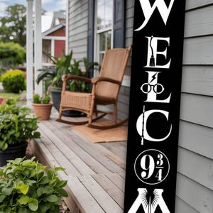 May include: A black wooden welcome sign with white lettering, featuring a bird, lightning bolt, glasses, wand, and the numbers 9 3/4. The sign is displayed on a porch with a rocking chair and plants.