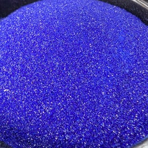 May include: A container filled with a large quantity of fine, vibrant royal blue sand. The granular texture of the sand is clearly visible, creating a dense and uniform surface within the circular black rim of the holding vessel.