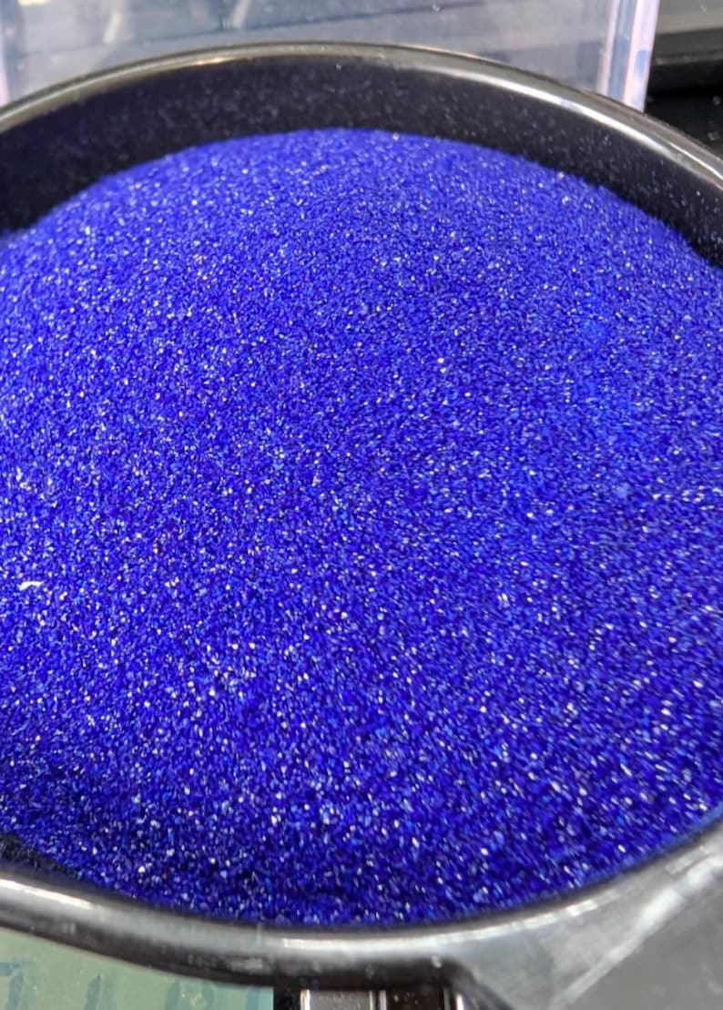 May include: A container filled with a large quantity of fine, vibrant royal blue sand. The granular texture of the sand is clearly visible, creating a dense and uniform surface within the circular black rim of the holding vessel.