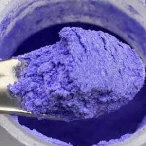 May include: A metallic purple pigment powder held on a small silver metal spatula. The fine, vibrant violet powder is shown in a concentrated heap on the tool, with a portion of the powder remaining inside a white circular container.