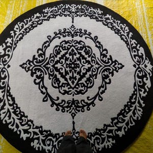 May include: A circular rug featuring a high-contrast black and white color scheme. The design consists of a central diamond-shaped motif surrounded by intricate, symmetrical scrollwork patterns that extend toward the outer black border.