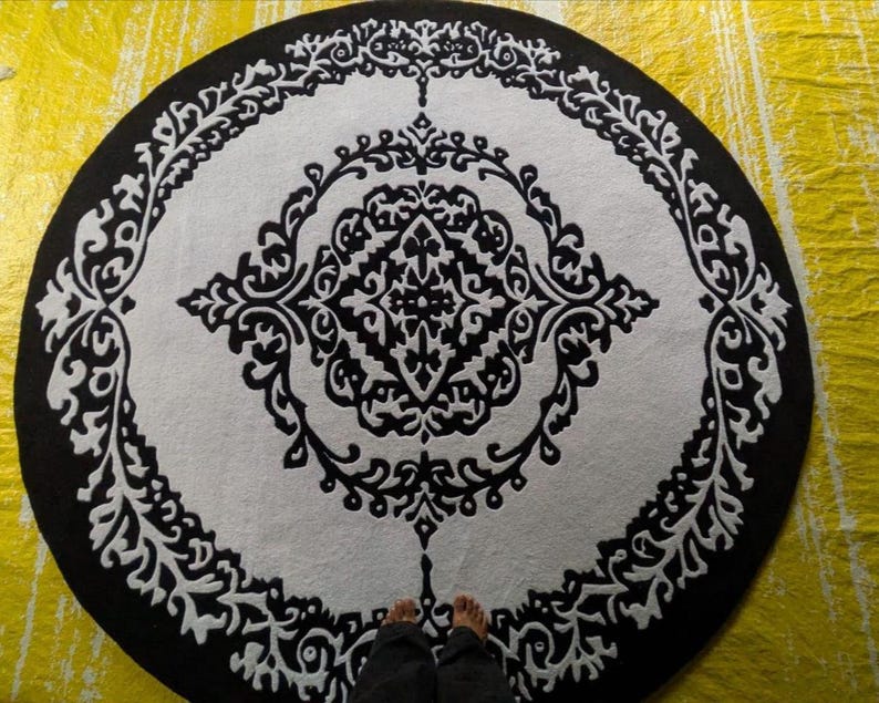 May include: A circular rug featuring a high-contrast black and white color scheme. The design consists of a central diamond-shaped motif surrounded by intricate, symmetrical scrollwork patterns that extend toward the outer black border.