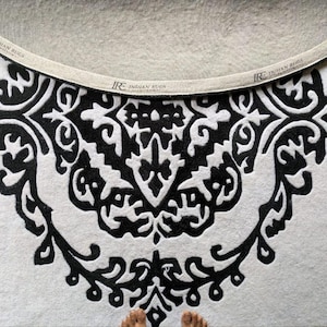 May include: A circular rug featuring a high-contrast black and white pattern with intricate symmetrical scrollwork. The edge of the rug is finished with a beige fabric binding tape printed with the text IRE INDIAN RUGS at regular intervals.