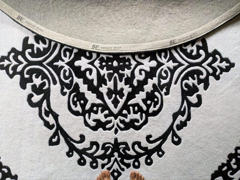 May include: A circular rug featuring a high-contrast black and white pattern with intricate symmetrical scrollwork. The edge of the rug is finished with a beige fabric binding tape printed with the text IRE INDIAN RUGS at regular intervals.
