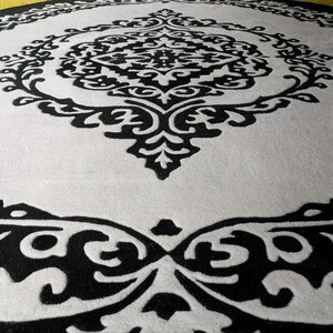 May include: A circular rug featuring a high-contrast black and white color scheme. The design consists of a central black ornate medallion pattern surrounded by a white field, which is enclosed by a wide black border with intricate white scrollwork.