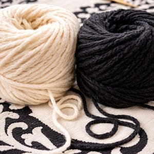May include: Two round balls of thick yarn, one in cream white and one in solid black, are positioned side by side. A pair of thin, metallic gold knitting needles rests diagonally in the background behind the yarn balls.