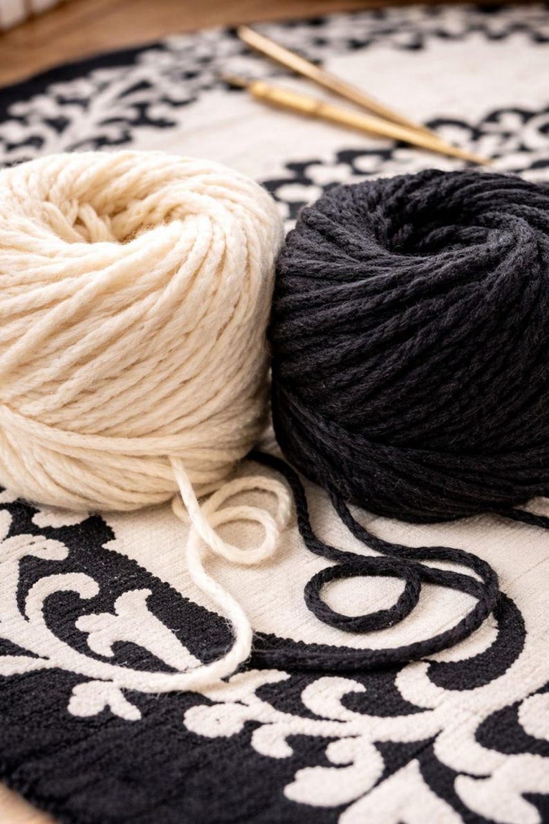 May include: Two round balls of thick yarn, one in cream white and one in solid black, are positioned side by side. A pair of thin, metallic gold knitting needles rests diagonally in the background behind the yarn balls.