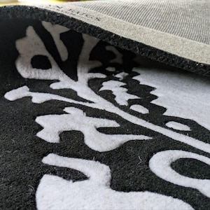 May include: A black and white rug with a folded corner revealing a gray textured backing. The rug features a prominent white botanical or leaf-like pattern set against a solid black background. The text "AN RUGS" is visible on the gray backing.