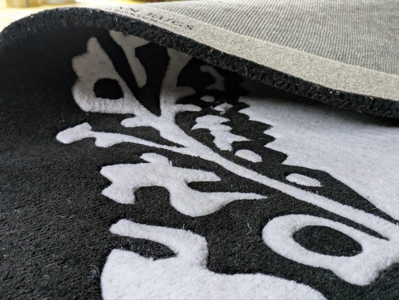 May include: A black and white rug with a folded corner revealing a gray textured backing. The rug features a prominent white botanical or leaf-like pattern set against a solid black background. The text "AN RUGS" is visible on the gray backing.