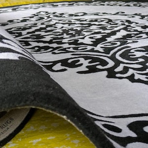 May include: A black and white area rug featuring a large, intricate, symmetrical damask-style pattern. The rug is partially folded over, revealing a thick, dark pile and a fabric label on the underside that reads INDIAN RUGS EMPORIUM.
