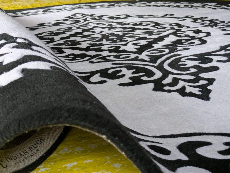 May include: A black and white area rug featuring a large, intricate, symmetrical damask-style pattern. The rug is partially folded over, revealing a thick, dark pile and a fabric label on the underside that reads INDIAN RUGS EMPORIUM.