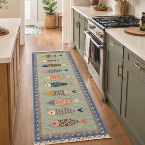 Pode incluir: Um tapete passadeira retangular longo com fundo verde claro e um padrão repetitivo de peixes estilizados em várias cores e designs, emoldurado por uma borda azul escura com padrão geométrico e finalizado com franjas brancas nas extremidades.