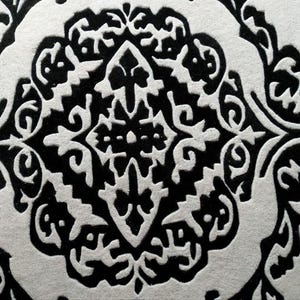 May include: A high-contrast black and white surface featuring a repeating damask-style pattern. The design consists of intricate, symmetrical, and ornate shapes with bold black lines set against a textured, off-white background material.