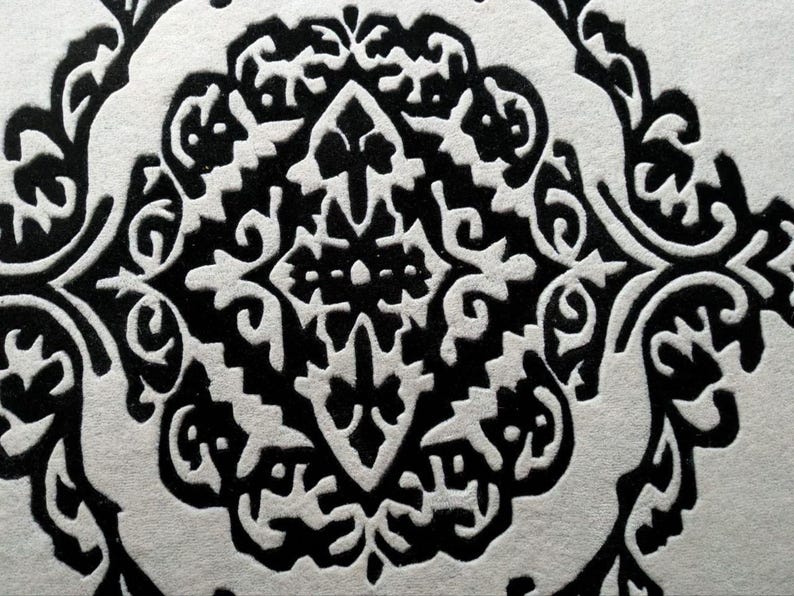 May include: A high-contrast black and white surface featuring a repeating damask-style pattern. The design consists of intricate, symmetrical, and ornate shapes with bold black lines set against a textured, off-white background material.