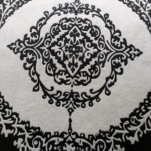 May include: A circular rug featuring a high-contrast black and white pattern. The design consists of a central diamond-shaped motif surrounded by intricate, symmetrical scrollwork and floral-inspired filigree elements arranged in concentric rings.