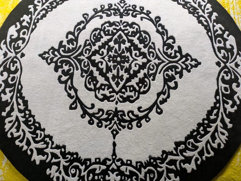 May include: A circular rug featuring a high-contrast black and white pattern. The design consists of a central diamond-shaped motif surrounded by intricate, symmetrical scrollwork and floral-inspired filigree elements arranged in concentric rings.