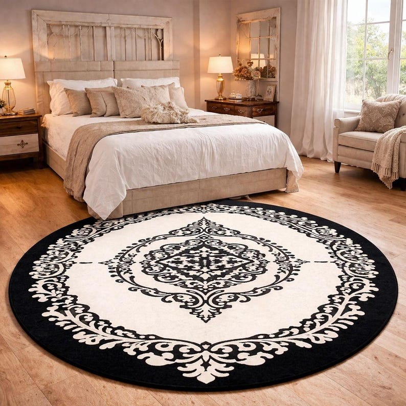 May include: A large circular area rug featuring a cream-colored center and a wide black border. The rug displays a symmetrical, intricate black damask-style pattern that radiates from the center toward the outer edge, creating a bold contrast.
