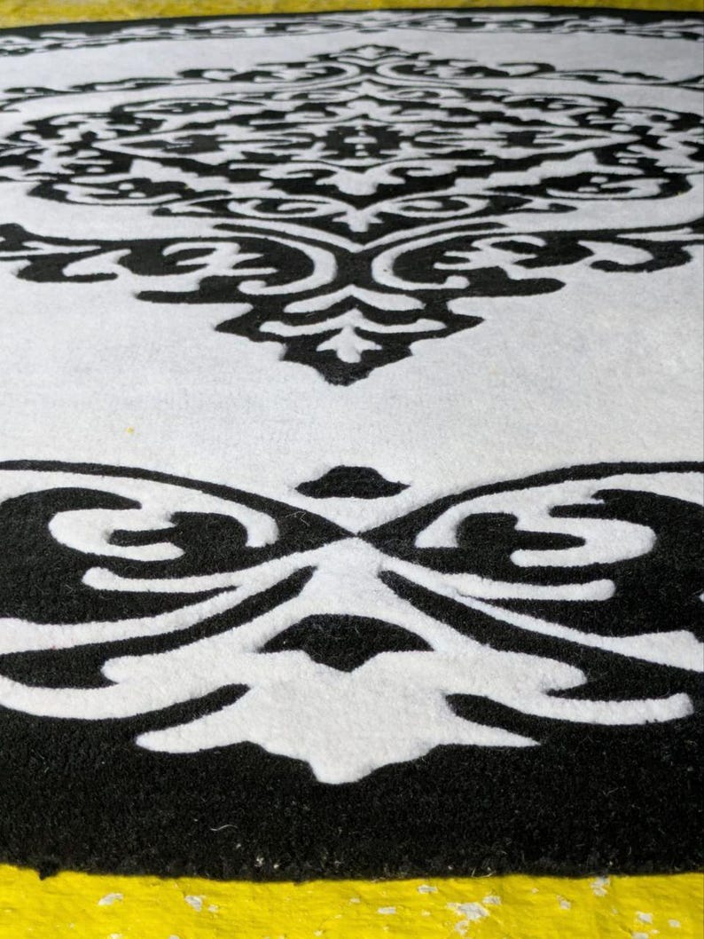 May include: A circular rug featuring a high-contrast black and white damask pattern. The design consists of intricate, symmetrical scrollwork and floral-inspired motifs that repeat across the surface, creating a bold graphic visual effect.