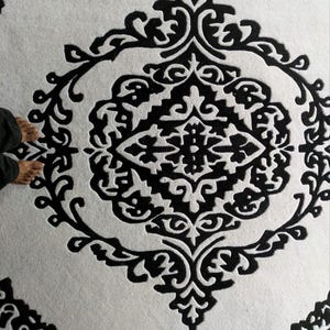 May include: A circular rug featuring a high-contrast black and white color scheme. The design consists of a central diamond-shaped motif surrounded by intricate, symmetrical scrollwork and floral-inspired patterns within a circular border.