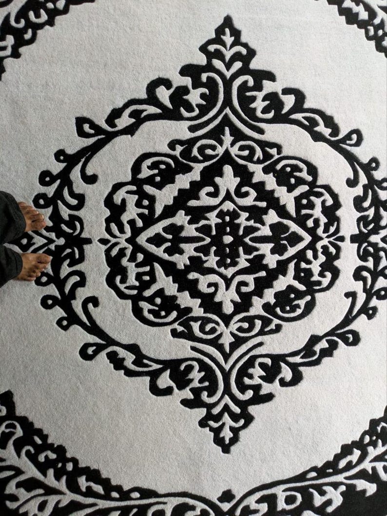 May include: A circular rug featuring a high-contrast black and white color scheme. The design consists of a central diamond-shaped motif surrounded by intricate, symmetrical scrollwork and floral-inspired patterns within a circular border.