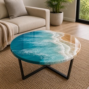 Op de afbeelding: Ronde salontafel met een harsblad dat een oceaan golf ontwerp weergeeft. De tafel heeft een zwart metalen onderstel en staat op een geweven vloerkleed. De oceaan-thema tafel heeft tinten blauw, wit en bruin. De diameter is ongeveer 90 cm.