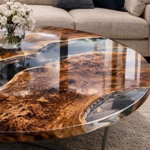 May include: A unique coffee table with a live edge wood design and a clear epoxy resin river. The table has a polished finish, showcasing the natural wood grain and a metallic edge. A glass vase with white flowers sits on the table.