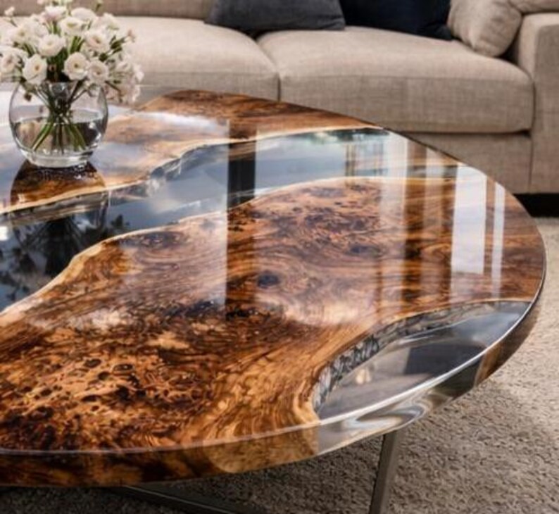 May include: A unique coffee table with a live edge wood design and a clear epoxy resin river. The table has a polished finish, showcasing the natural wood grain and a metallic edge. A glass vase with white flowers sits on the table.