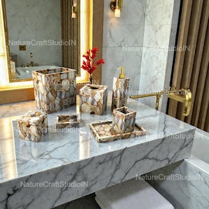 Puede incluir: Un juego de baño que incluye un cesto de basura, una caja de pañuelos, un dispensador de jabón, un vaso, una bandeja y un jabonera. El juego está hecho de un mosaico de piedras marrones, beige y crema. Los artículos están colocados sobre una encimera de mármol.