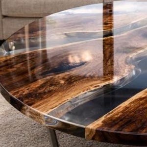 May include: A round coffee table with a dark wood grain pattern and a clear epoxy resin river. The table has a polished finish and sits on metal legs. The wood has a rich, brown color with lighter streaks.