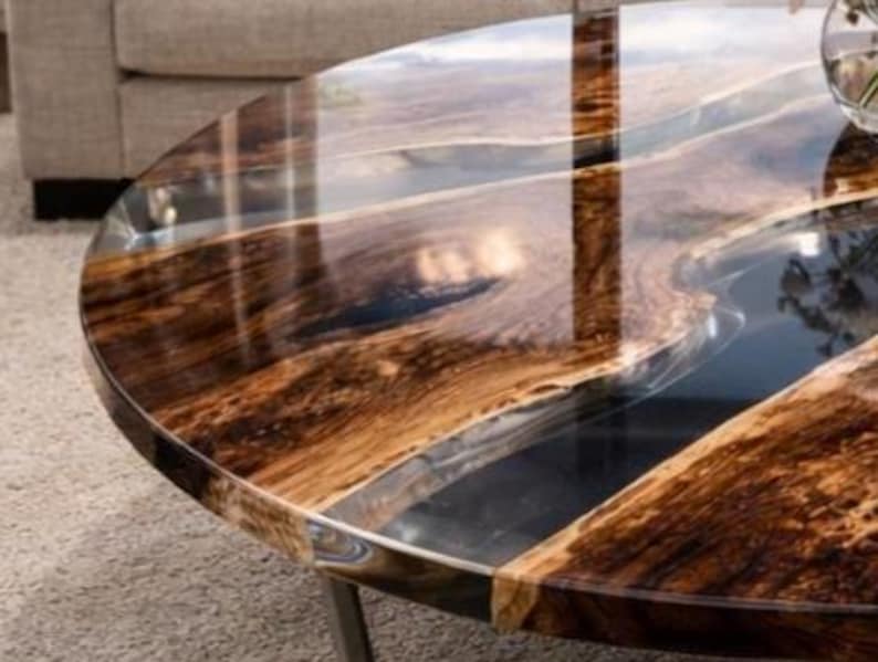 May include: A round coffee table with a dark wood grain pattern and a clear epoxy resin river. The table has a polished finish and sits on metal legs. The wood has a rich, brown color with lighter streaks.