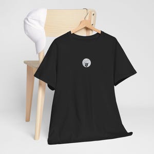 May include: Black t-shirt with a small, circular graphic of two figures silhouetted against a full moon. A white baseball cap rests on a wooden chair. The shirt is hanging on a wooden hanger.