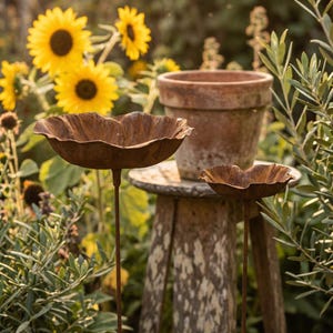 Puede incluir: Dos bebederos para p&aacute;jaros de metal oxidado con forma de flor sobre finos tallos, colocados en un jard&iacute;n con girasoles y vegetaci&oacute;n. Una maceta de terracota se encuentra sobre un taburete de madera desgastada. Los bebederos tienen un dise&ntilde;o texturizado en forma de p&eacute;talo.
