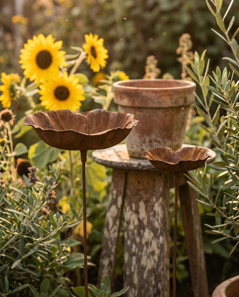 Puede incluir: Dos bebederos para p&aacute;jaros de metal oxidado con forma de flor sobre finos tallos, colocados en un jard&iacute;n con girasoles y vegetaci&oacute;n. Una maceta de terracota se encuentra sobre un taburete de madera desgastada. Los bebederos tienen un dise&ntilde;o texturizado en forma de p&eacute;talo.