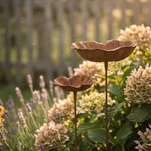 Puede incluir: Dos bebederos decorativos de metal con aspecto envejecido y dise&ntilde;o floral. Los bebederos son marrones y se alzan sobre finos postes. Est&aacute;n situados en un jard&iacute;n con arbustos de lavanda e hortensias, con una valla de madera al fondo.