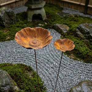 Puede incluir: Dos esculturas de flores de metal oxidado con un acabado texturizado. Las flores est&aacute;n sobre finos tallos, colocadas en un jard&iacute;n zen con grava gris y musgo verde. Una linterna de piedra y una valla de bamb&uacute; est&aacute;n en el fondo.